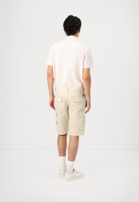 Pantaloni cargo beige con molte tasche laterali, abbinati a una camicia rosa chiaro a maniche corte, visti da dietro. Calze bianche e scarpe da ginnastica.