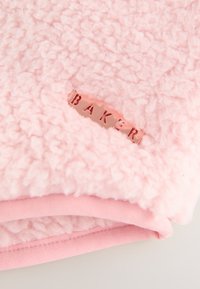 Pink tekstureret fleece-stof med et lille metallisk rosaguld-mærke med mærkenavnet "Baker" syet ind i det.