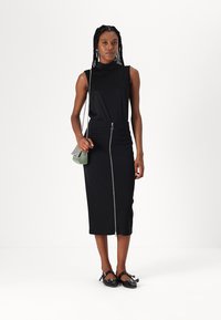 Top negru fără mâneci asociat cu o fustă tip creion neagră, ajustată, cu fermoar frontal. Accesoriile includ o geantă mică cu carouri verzi și pantofi joși.