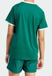 Zelené bavlněné tričko s bílým lemem, s kulatým výstřihem a krátkými rukávy, v kombinaci se zelenými šortkami. Pohled zezadu s hladkou texturou.