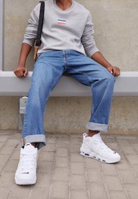 Osoba siedząca na ławce, ubrana w szary sweatshirt Levi's, niebieskie dżinsy z podwiniętymi nogawkami, białe sneakersy Nike oraz czarną torbę na ramię.