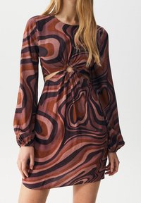 Nainen, jolla on päällä pitkähihainen mini-mekko, jossa on ruosteen, ruskean ja mustan swirl-kuvioinnin sekä pyöreä läppä yksityiskohtana vyötäröllä.