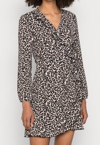 Kobieta w długiej, leopardo wzorzystej sukience z długim rękawem, z falbanami wzdłuż dekoltu, talii i dolnej krawędzi.