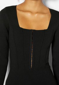 Body in maglia nera con scollatura quadrata, maniche lunghe, dettagli di cuciture verticali e una fila centrale di chiusure a scatto.