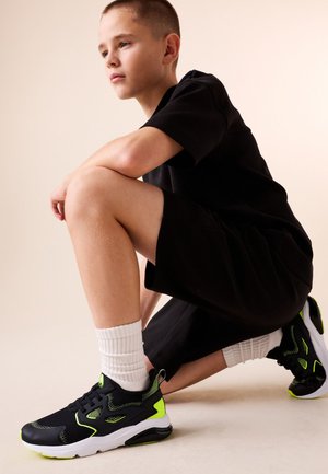 Jongen in zwarte outfit hurkend, met zwarte en neon groene sneakers met witte sokken, nadenkend naar de zijkant kijkend tegen een lichte achtergrond.