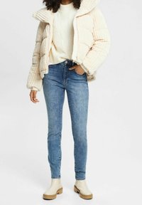 Pufferjack in lichtroze met geribbelde textuur, gedragen over een crèmekleurige gebreide trui en blauwe slim-fit jeans, gecombineerd met beige chunky-sole laarzen.
