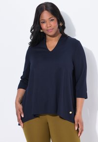 Tummansininen V-pääntie-tunika, jossa on kolmen neljänneksen hihat, aaltoileva muotoilu, hieman pidempi takapohja ja pieni kultainen yksityiskohta alaosassa.