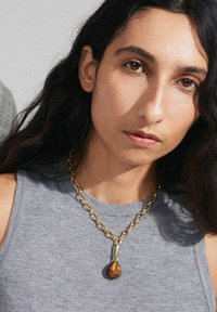 Gold chain necklace with a teardrop-shaped tiger's eye pendant. Model wears a gray sleeveless top, showcasing the jewelry's details.