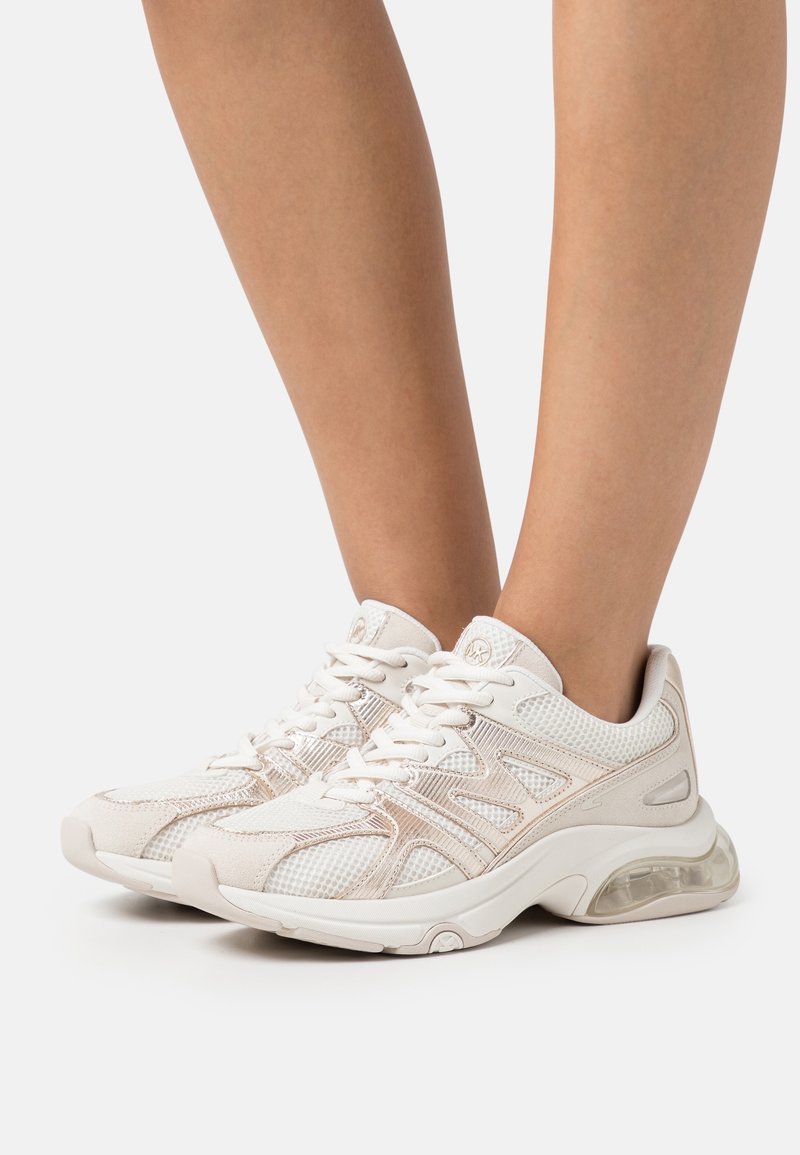 Pieds portant des baskets en mesh blanc et beige avec des semelles épaisses et un coussin d'air visible, présentés sur un fond clair uni.