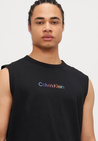 Svart ermeløs T-skjorte laget av bomull, med en gradient "Calvin Klein"-logo i pastelldfarger over brystet.