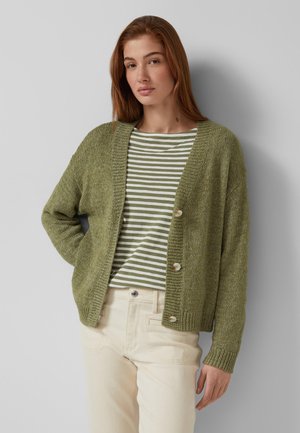Young woman wearing an olive green cardigan over a striped green and white shirt with cream pants, posing against a light gray background.