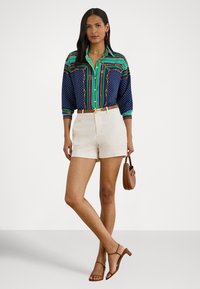 Kvinne iført en mønstrete grønn og marineblå bluse, beige shorts, brune sandaler og håndveske, stående foran en ensfarget bakgrunn.