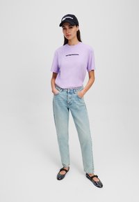 Lila T-shirt met korte mouwen en zwarte tekst, lichtblauwe high-waisted jeans en zwarte flats met stippen, gecombineerd met een zwarte pet.