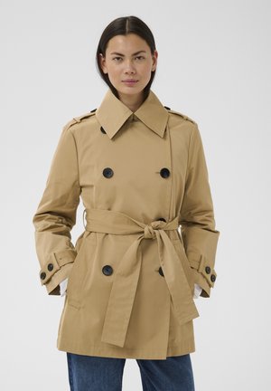 Naine, kellel on seljas beež kahekordse nööpimisega vihmamantel, millel on seotud vöö, mustad nupud ja sinised teksad, tavalise valge tausta ees.