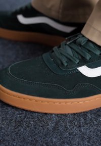 Sapatilha verde em camurça com sola de borracha castanha, apresentando detalhes em branco e um design clássico de atacadores. Parte superior texturizada com costuras decorativas.