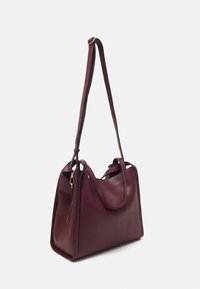 Sac tote en cuir bordeaux avec une texture lisse, double poignées, sangle ajustable et détail de fermeture éclair dorée sur le côté.
