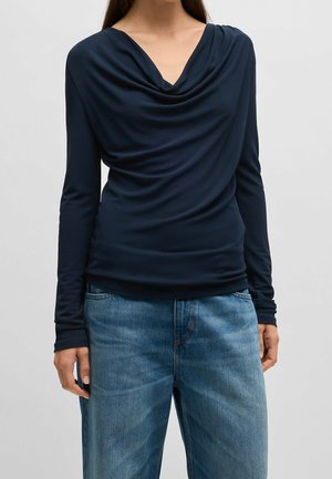 Kvinde iført navyblå bluse med lange ærmer og draperet halsudskæring, kombineret med blå denimjeans, stående foran en ensfarvet baggrund.