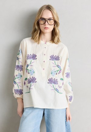 Junge Frau mit blondem Haar und Brille, die eine cremefarbene Bluse mit violetter Blumengestickerei und hellblaue Jeans trägt, vor einem neutralen Hintergrund stehend.