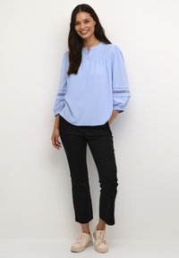 Blusa celeste con scollatura a bottoni, parte anteriore plissettata e dettagli in pizzo sulle maniche, abbinata a pantaloni neri a tre quarti e sneakers beige.