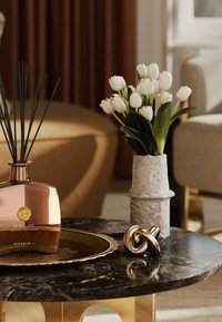 Reed diffuser i en rosegullfarget glassflaske med svarte staver, ved siden av en grå keramikkvase med hvite tulipaner og et brett med gullaccent på et svart marmorbord.