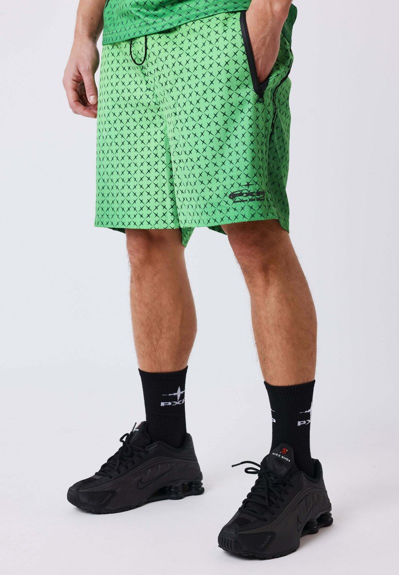 Personne portant un short vert à motifs, des chaussettes noires de marque et des baskets Nike Shox noires, debout devant un fond clair uni.