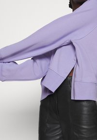 Sudadera de color lila con mangas largas, puños acanalados, aberturas laterales y un diseño corto. La tela parece suave con una textura lisa.