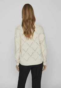 Kremowy sweter dziergany z rombowymi wycięciami, z prążkowanymi mankietami i dołem. Noszony na czarnych spodniach, prezentujący luźny krój i teksturowany wzór.