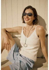 White knitted tank top with thin straps, light blue shorts, and a circular pendant necklace. Model wearing black oval sunglasses.