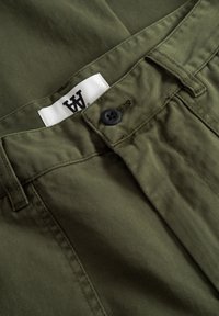 Bombažne hlače olivne zelene barve z gladko teksturo, opremljene s črnim gumbom in belo etiketo s črnim logotipom blizu pasu.