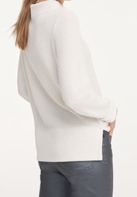 Biały, teksturowany sweter z wysokim dekoltem i długimi rękawami, w połączeniu z ciemnoszarymi dżinsami. Sweter ma luźny krój i boczne rozcięcia.