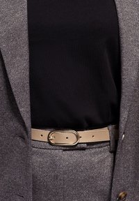 Grauer Fischgrät-Blazer mit schwarzem Oberteil darunter, in der Taille von einem beigen Gürtel mit ovaler goldener Schnalle geschnürt.