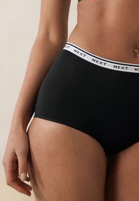 Sous-vêtements noirs avec ceinture élastique blanche portant le logo "NEXT", en coton, de coupe taille haute, texture lisse et forme ajustée.