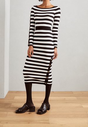 Mujer con vestido midi de manga larga a rayas blancas y negras con botones dorados, medias negras y sandalias de tacón negras con puntera abierta.