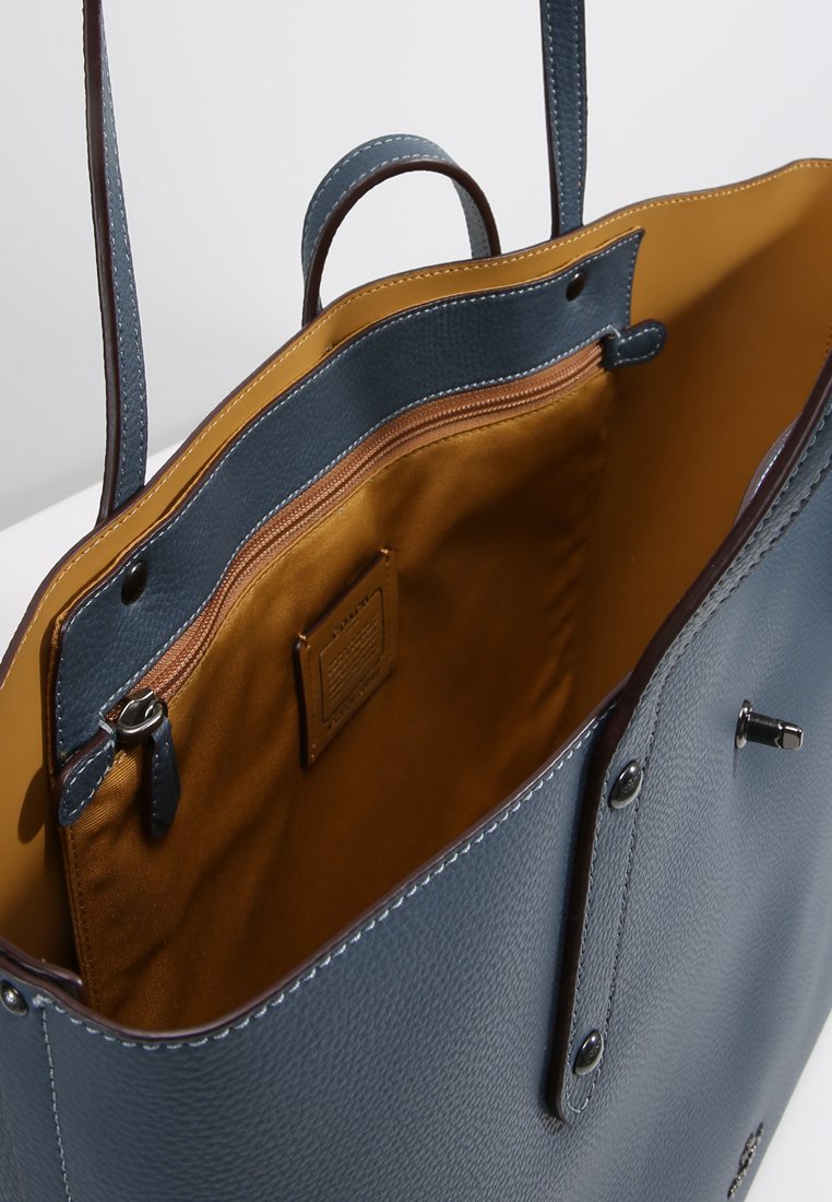 Bolso tote de cuero azul con un acabado texturizado, que presenta un forro de tela marrón, un bolsillo con cremallera y detalles de costura en la parte superior. Diseño abierto.