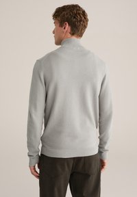 Pulover tricotat gri deschis cu guler inalt, manșete și tiv tip dungi. Model texturat pe toată suprafața. Purtat cu pantaloni verde închis.