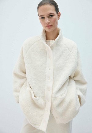 Mujer con el pelo peinado hacia atrás, que lleva una chaqueta de forro polar blanca con botones y tiene las manos en los bolsillos, frente a un fondo claro y liso.