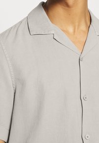 Camicia grigia chiara a maniche corte in un tessuto leggero, con colletto alla marinara, chiusura con bottoni e superficie texturizzata sottile.