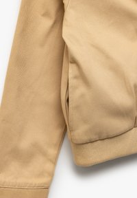 Giacca in cotone beige con polsini a costina, caratterizzata da una trama liscia e dettagli con tasche laterali. Linee pulite con un design moderno e su misura.