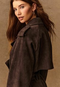 Trench coat in suede marrone con una vestibilità rilassata, mostrine sulle spalle e vita con cintura. Presenta un colletto ampio e dettagli di cucitura in vista.