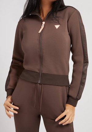 Frau trägt braune Jacke mit Reißverschluss und Kordelzughosen mit gerippten Bündchen und dezentem Logo auf Ärmeln und Brust.