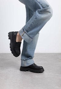 Zapatos negros de cuero tipo deslizante con una suela gruesa y texturizada, combinados con jeans azul claro con dobladillo, mostrando un diseño elevado y grueso.
