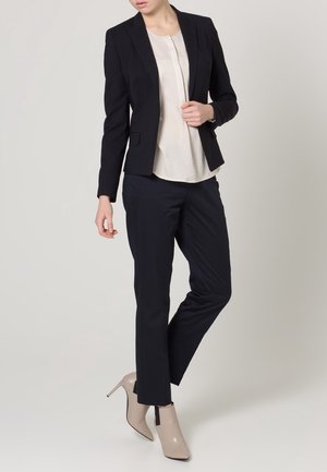 Mujer con blazer negro, blusa blanca, pantalones negros y botines beige de tacón alto, de pie con una pierna ligeramente levantada.