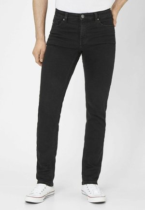 Mand iført slim-fit sorte jeans og hvide sneakers, stående foran en ensartet lys baggrund.
