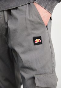 Hand i fickan på gråa cargobyxor med sydd logotypmärke som har en orange och röd halvcirkel på svart bakgrund.