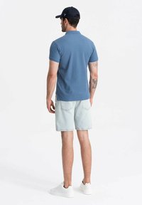 Modrá polo tričko vyrobené z bavlny, v kombinaci s světlými džínovými šortkami. Bílé tenisky doplňují outfit. Viditelné tetování na levém rameni.