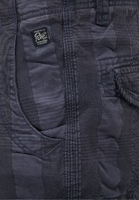 Tmavě modrá, texturovaná tkanina s jemným pruhovaným vzorem. Obsahuje kapsu a značkovou etiketu našitou na boku.