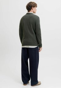 Mørkegrøn pullover med ribbet kant, båret over en hvidstribet skjorte, parret med løstsiddende navy bukser og beige sko.