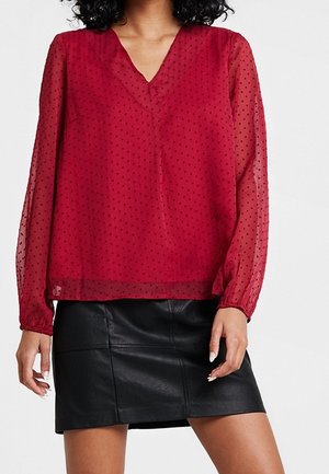 Vrouw draagt een doorschijnende rode blouse met lange mouwen met kleine stippen en een zwarte leren minirok, staand tegen een witte achtergrond.