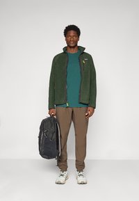 Green fleece jacket with black zipper and yellow zip pull, teal shirt, brown pants, black backpack, gray hiking shoes, standing against a light background.
