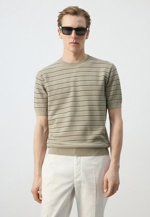 Hombre con gafas de sol negras, suéter de punto beige de manga corta con finas rayas negras y pantalones blancos, de pie frente a un fondo claro y liso.